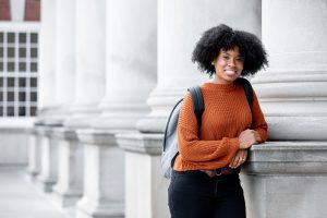 Syracuse University student Taylor John wearing an orange sweater and gray backpack standing near columns cascading in background.